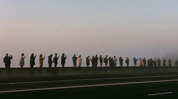 a line of spectators along a freeway barrier at dawn holding phones up, fog below, pale dawn grey-white, soft even light, a single clay-orange #D97757 accent, fine grain, calm, no text, no letters, no logos