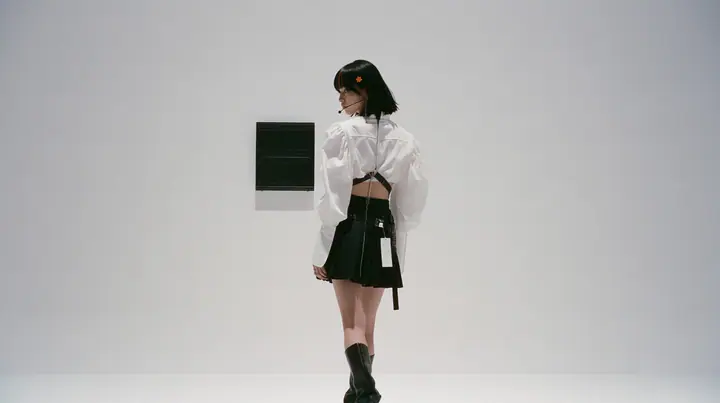 wide shot of a small black split-flap board with blank flaps alone on a seamless white paper wall, a young Caucasian American woman with pale skin and light freckles, a glossy black blunt jaw-length bob with heavy…