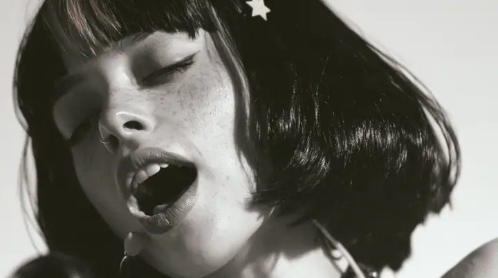 close-up of a young Caucasian American woman with pale skin and light freckles, a glossy black blunt jaw-length bob with heavy bangs and one clay-orange streak through the bangs, a small flat clay-orange…