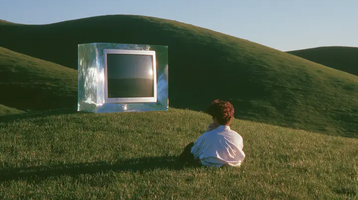 Y2K high fashion editorial photograph, 2001: a translucent ice-blue 2001 CRT computer monitor sitting alone on the crest of a smooth rolling bright-green grassy hill with soft curves, the grass short and velvety at…