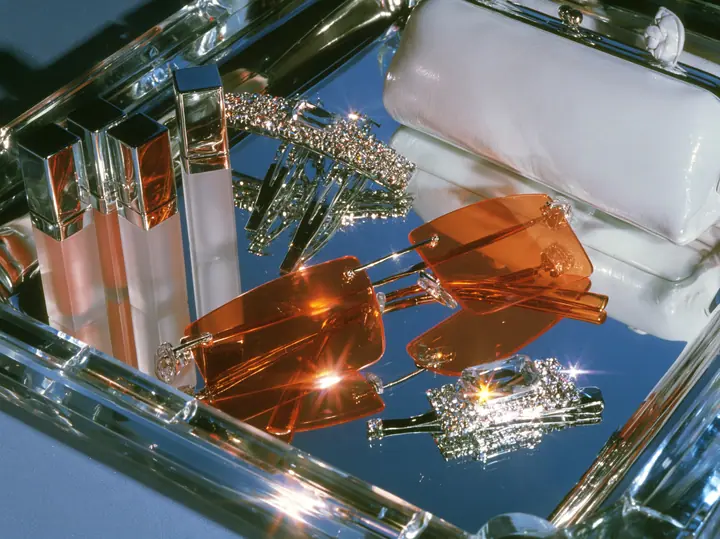 Y2K high fashion editorial photograph, 2001: a luxury beauty still life of frosted lip-gloss tubes, rimless orange-tinted shield sunglasses, rhinestone hair clips and a tiny white baguette bag on a mirrored tray,…